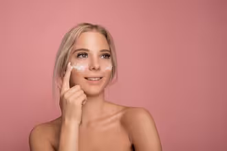 Pembe arka plan önünde, yüzüne krem süren genç bir kadın.