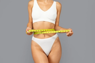 Gri arka plan önünde, beyaz iç çamaşırı giyen ve beline sarılı mezurayı tutan bir kadın.